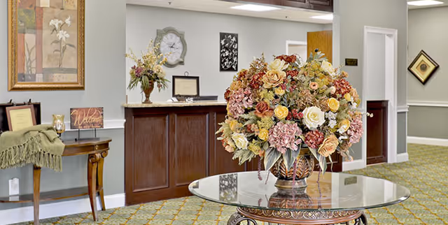 Interior view of a senior living facility lobby area featuring a round glass table with a large floral arrangement in the center. Behind the table is a wooden reception desk with decorative items including a clock, framed pictures, and a vase with flowers. The walls are painted light gray with white trim, and the floor is carpeted with a patterned design. There is a small wooden side table with a green throw and a welcome sign on the left side of the image.