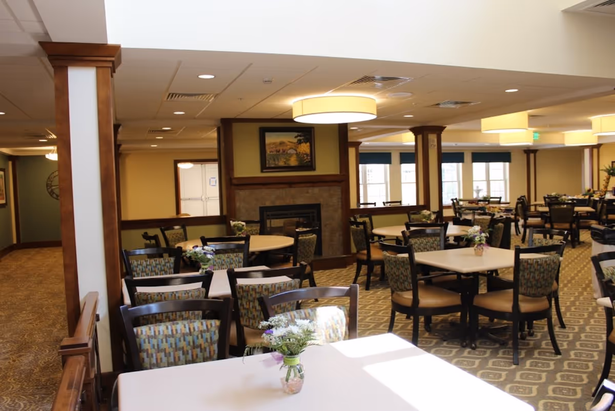 A spacious dining room in a retirement community with multiple tables and chairs arranged neatly. Each table has a small floral centerpiece. The room features warm lighting, a carpeted floor with a patterned design, wooden columns, and a fireplace with a painting above it. Large windows allow natural light to enter the space.