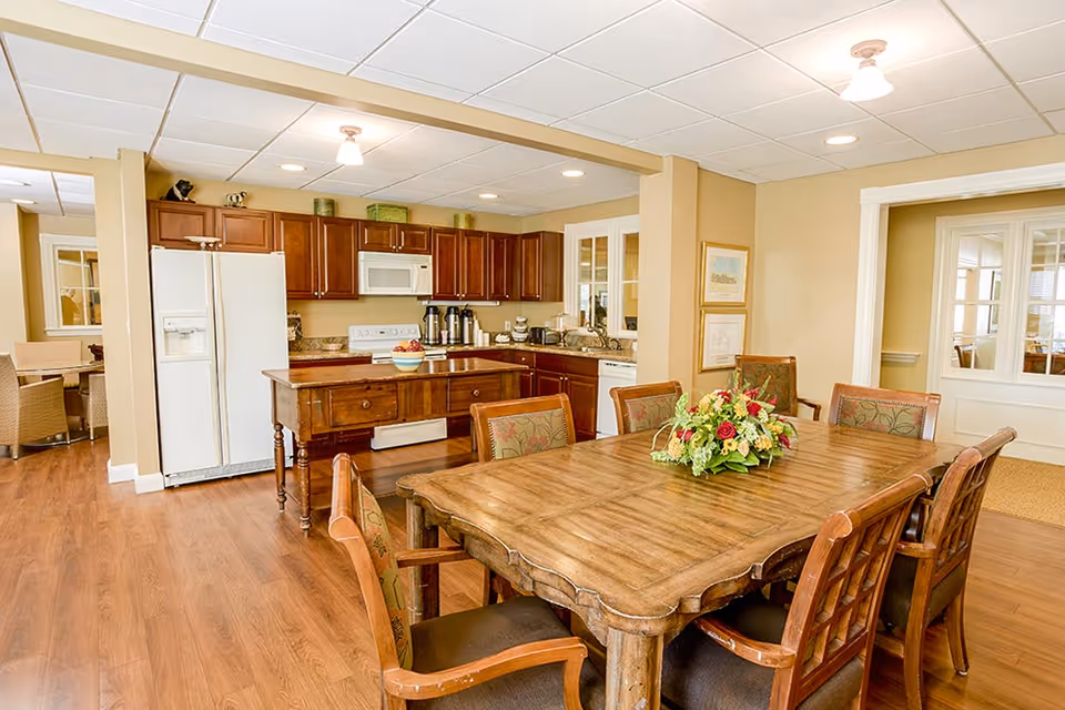 A warm and inviting kitchen and dining area with wooden floors and cabinetry. The kitchen features a white refrigerator, microwave, stove, and coffee station. In the foreground, there is a wooden dining table with six upholstered chairs and a floral centerpiece. The walls are painted beige, and there are framed pictures on the wall near a doorway with glass panels.