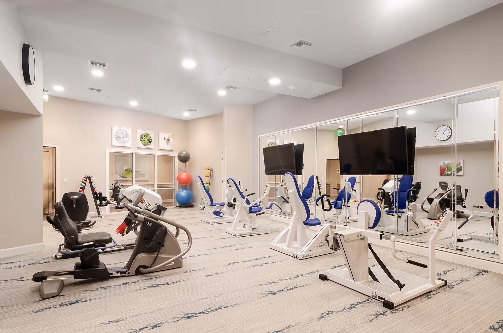 A well-lit fitness room with various exercise machines including recumbent bikes and strength training equipment. The room has a large wall mirror, a mounted TV, and colorful exercise balls stacked in the corner. The walls are light-colored with framed artwork, and the floor is carpeted with a subtle pattern.