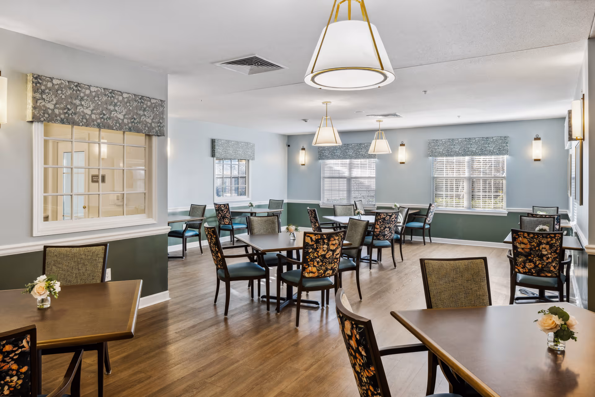 A bright dining room with multiple square tables and chairs arranged neatly. The chairs have floral patterned upholstery on the backrests. The room has light blue walls with a darker green wainscoting, wooden flooring, and several windows with floral valances allowing natural light to enter. Modern pendant lights hang from the ceiling, and small flower arrangements are placed on the tables.