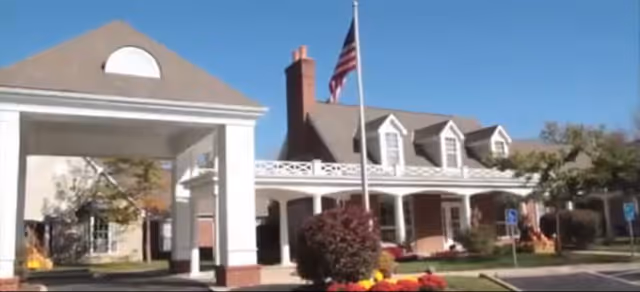 Exterior view of a senior living facility with a covered entrance, an American flag on a flagpole, and a building with dormer windows and a porch surrounded by landscaping and a parking area.