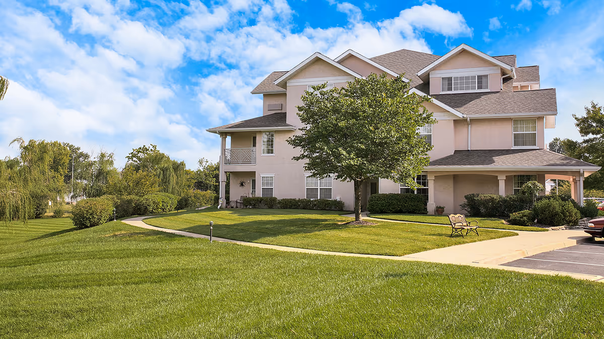 Exterior view of a multi-story residential building with beige walls and a gray roof, surrounded by a well-maintained lawn, trees, bushes, a sidewalk, and a bench under a blue sky with scattered clouds.
