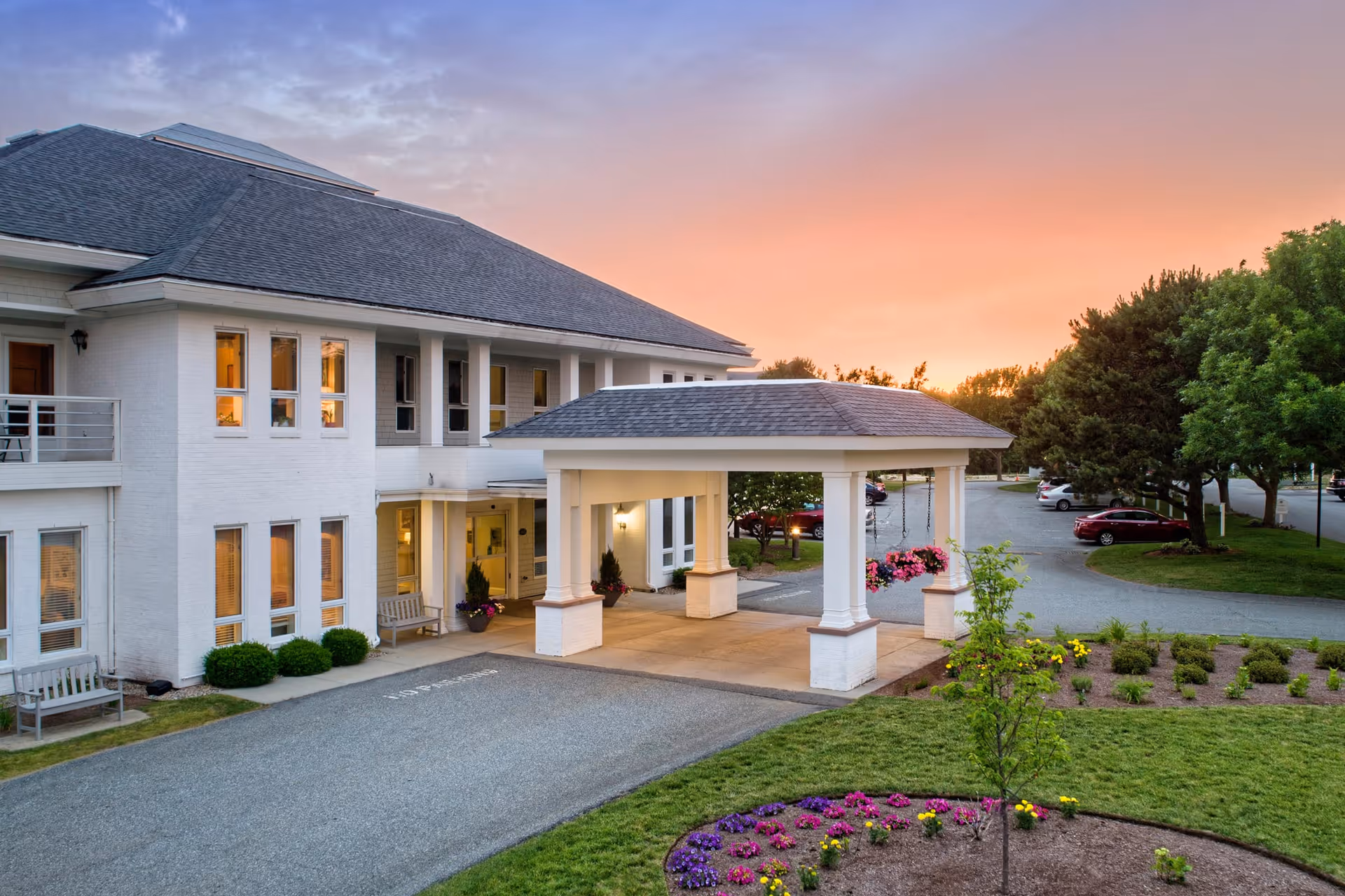 Exterior view of a senior living facility building at sunset with a covered entrance, landscaped garden with colorful flowers, parked cars, and trees surrounding the area.