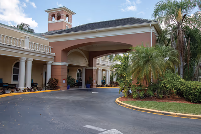 Covered porte-cochère entrance of a stucco senior living building with columns, palm trees, and a curved driveway.