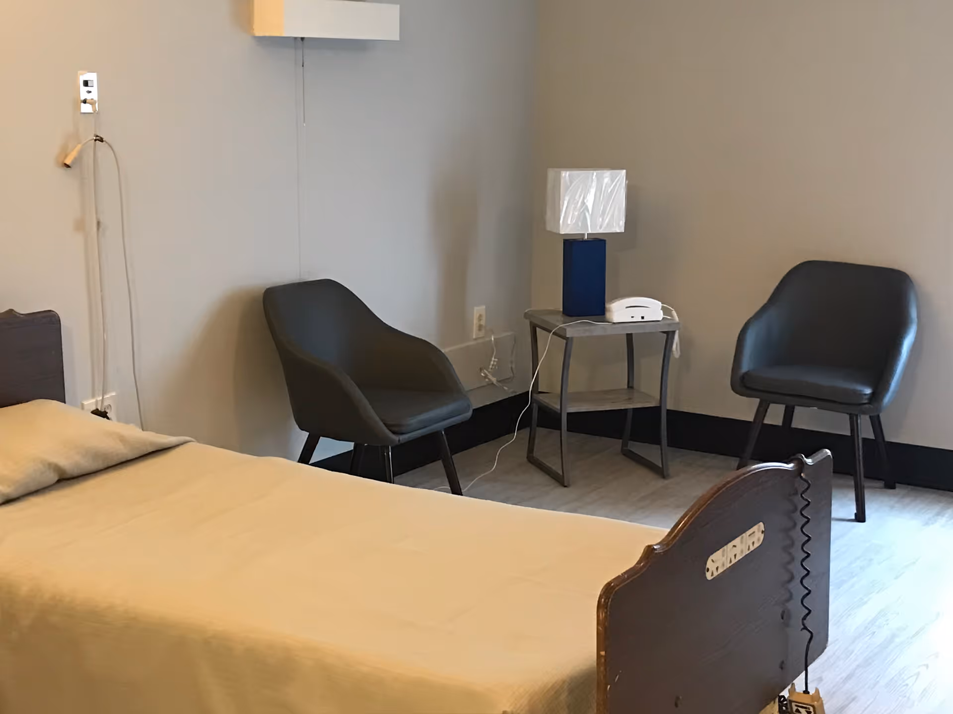 A simple room with a single bed covered in a beige blanket, two dark gray chairs, a small side table with a blue lamp and a white telephone, and light-colored walls and flooring.