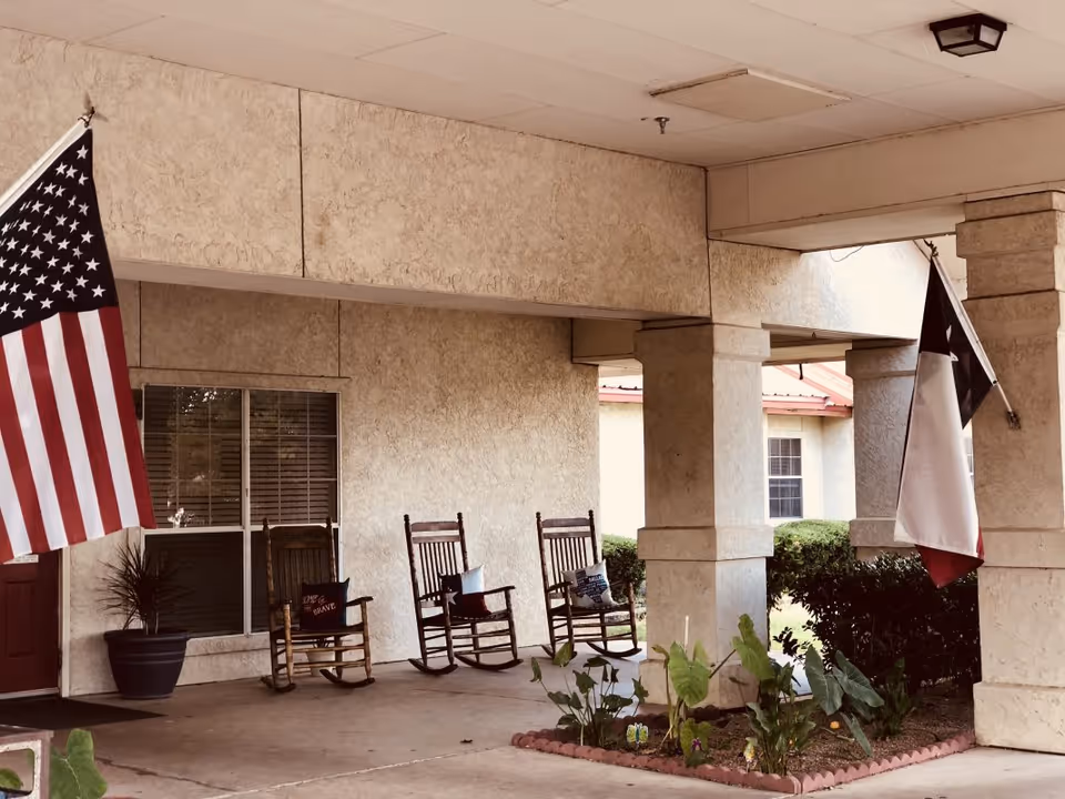 Covered front porch with rocking chairs, potted plants, and American and Texas flags at the entrance of a senior living facility.