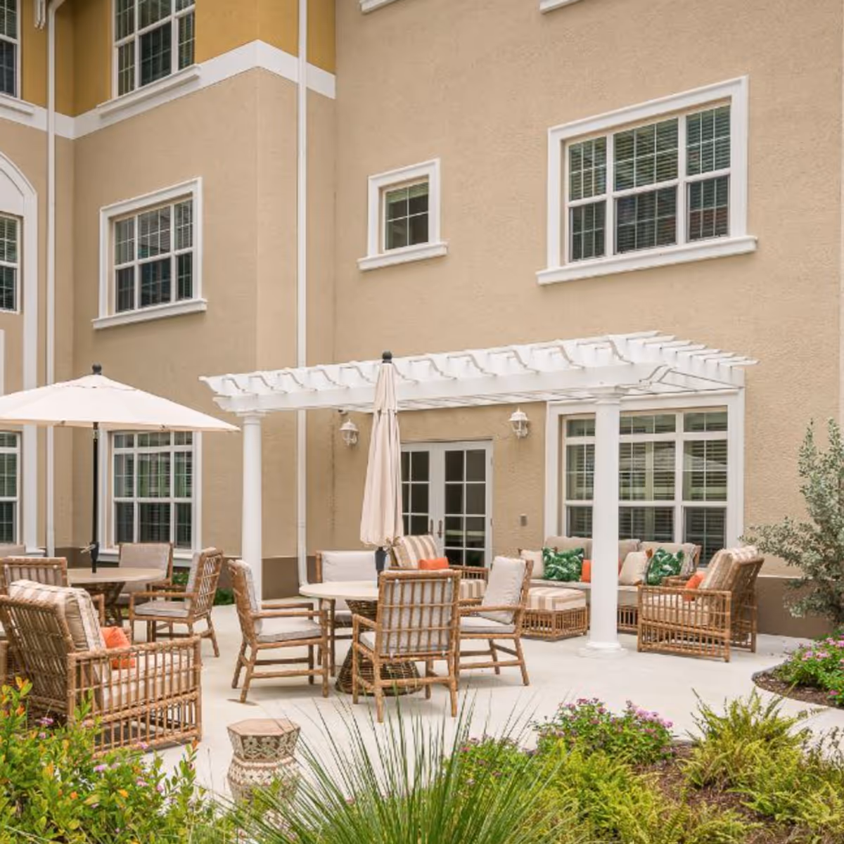 Outdoor patio with wicker chairs, tables, umbrellas, and a white pergola in front of a beige multi-story building.