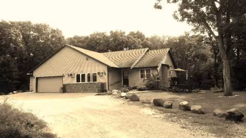 A single-story residential-style building with a wide driveway, stones, and mature trees.