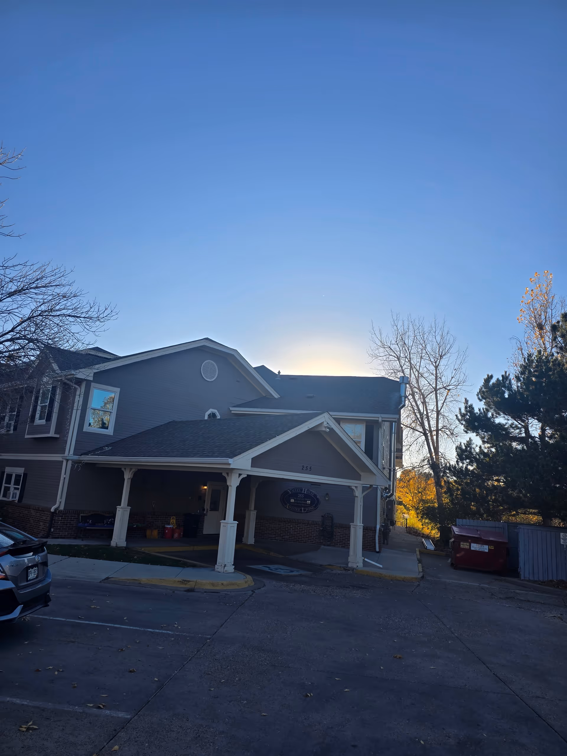 Exterior view of a two-story assisted living facility building with a covered entrance. The building has gray siding with white trim and a brick base. There are trees without leaves and a clear blue sky in the background. A car is partially visible in the parking area in front of the building.