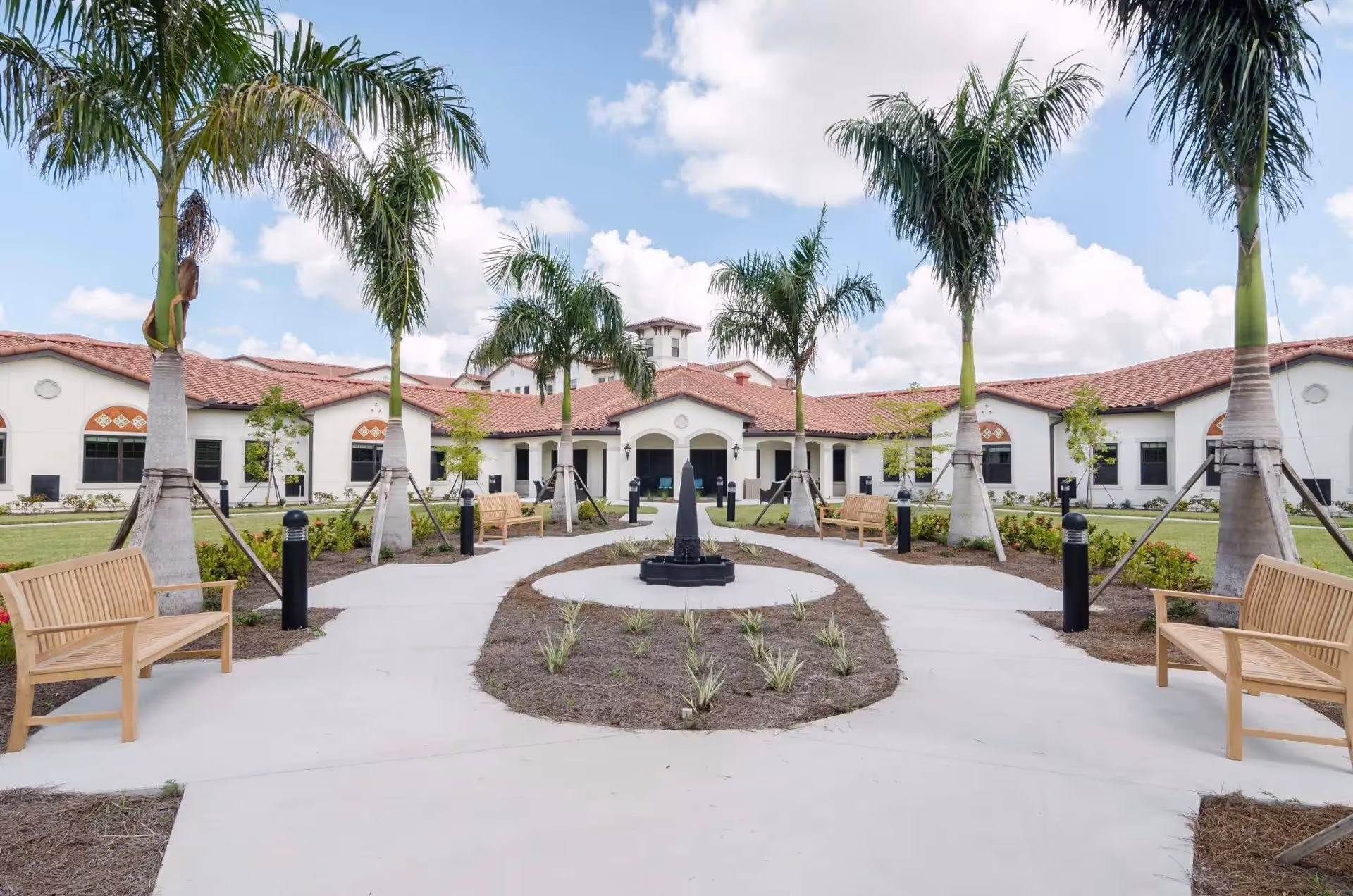 Outdoor courtyard area of a senior living facility with palm trees, benches, a central pathway, and a building with a red tile roof in the background under a partly cloudy sky.