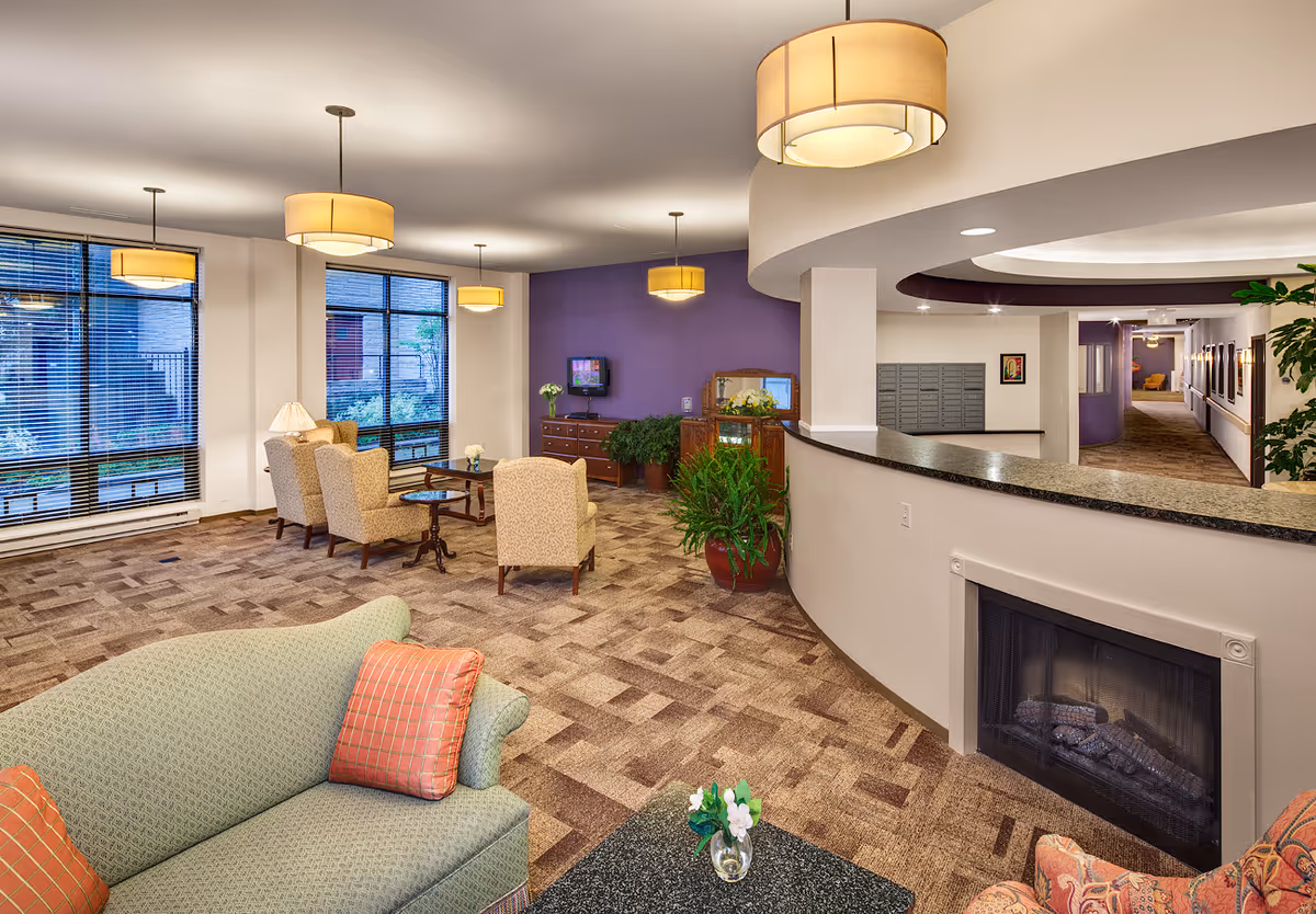 A spacious senior living facility common area with comfortable seating including armchairs and sofas, a fireplace, large windows with blinds, hanging ceiling lights, and various plants. The room has a purple accent wall with a small TV mounted on it and a hallway leading to other parts of the facility.