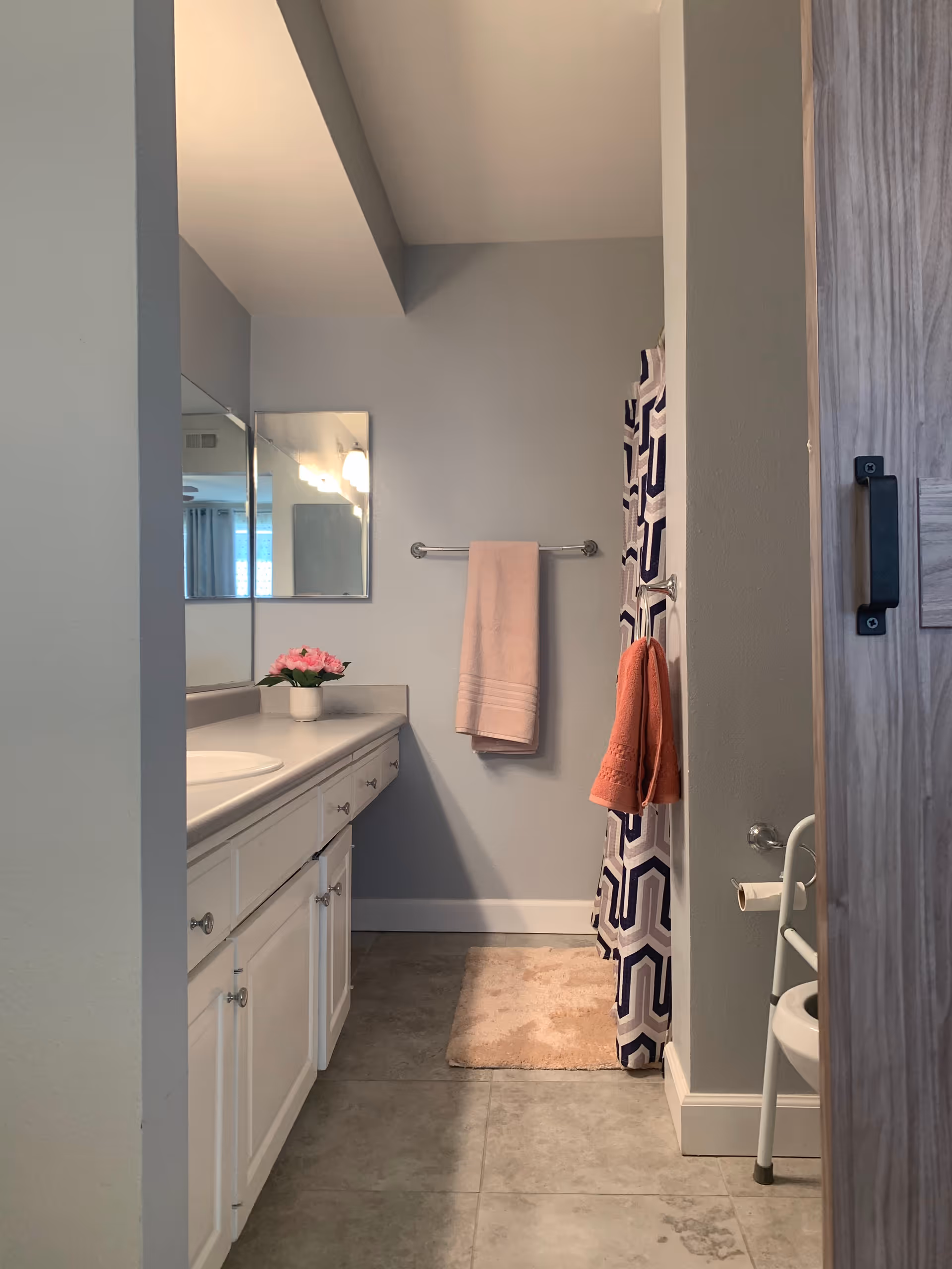 A clean bathroom with a white countertop and sink on the left, a mirror above the sink, a pink towel hanging on a towel rack on the back wall, a patterned shower curtain on the right, an orange hand towel hanging on a hook, a beige bath mat on the tiled floor, and a wooden door partially visible on the right side.