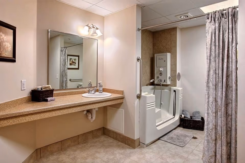 A senior living facility bathroom featuring a large mirror above a beige countertop with a sink. There is a walk-in bathtub with safety rails and a shower curtain. The floor is tiled, and there are baskets with towels near the bathtub. A framed picture hangs on the wall.
