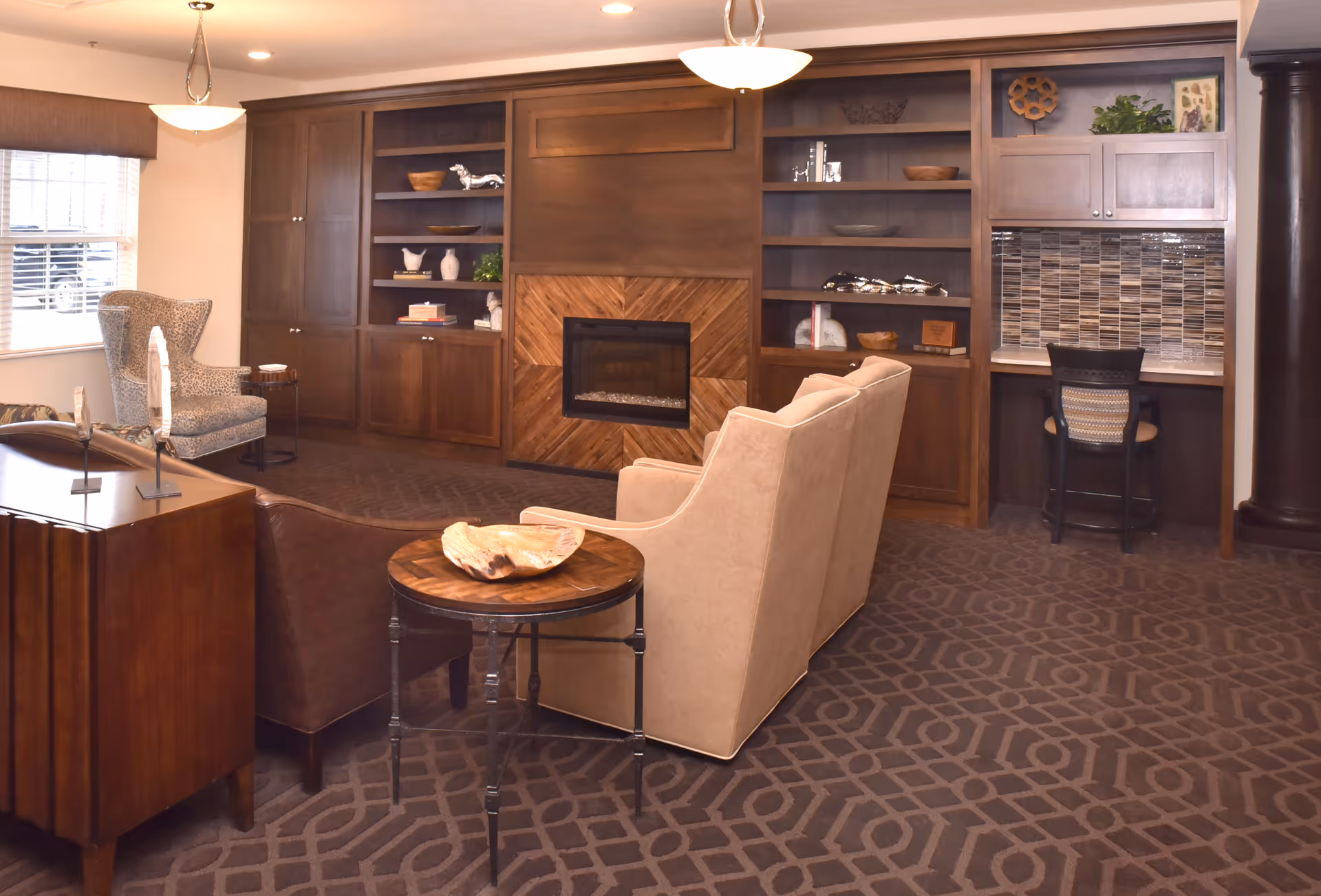 A cozy communal living room with sofas and armchairs facing a built-in wooden fireplace and shelving unit with a small desk area.