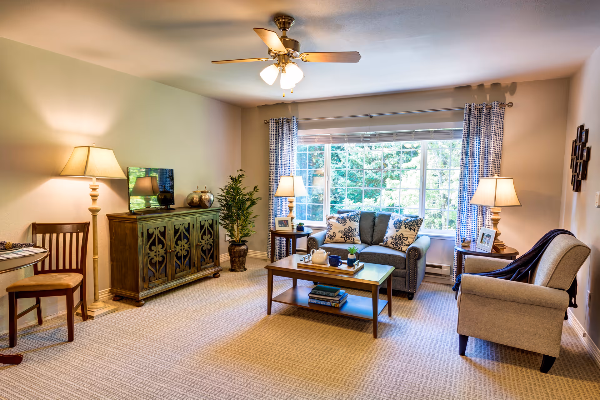 A cozy living room with a large window letting in natural light. The room features a gray loveseat with patterned cushions, a beige armchair with a dark throw, a wooden coffee table with books and a tea set, two side tables with lamps, a wooden cabinet with decorative items, a floor lamp, a small round table with a chair, and a ceiling fan with lights. The walls are light-colored and the floor is carpeted.