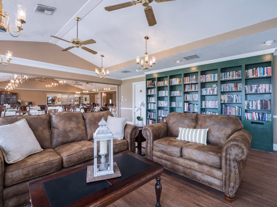Lounge area with two brown leather sofas, a coffee table, and a large bookshelf wall with a dining area visible in the background.