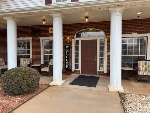 Brick building entrance with white columns, a brown door, welcoming sign, and porch seating.