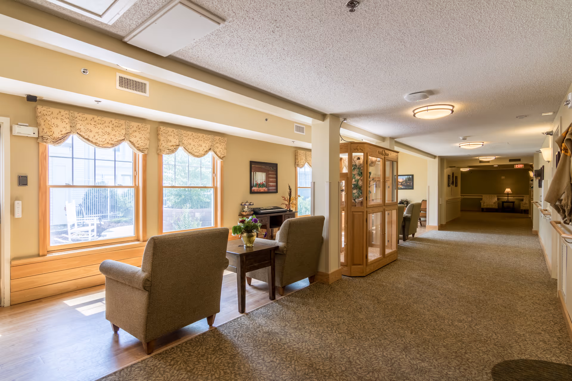 Sunlit senior living common area with armchairs and small tables along a carpeted hallway and large windows.