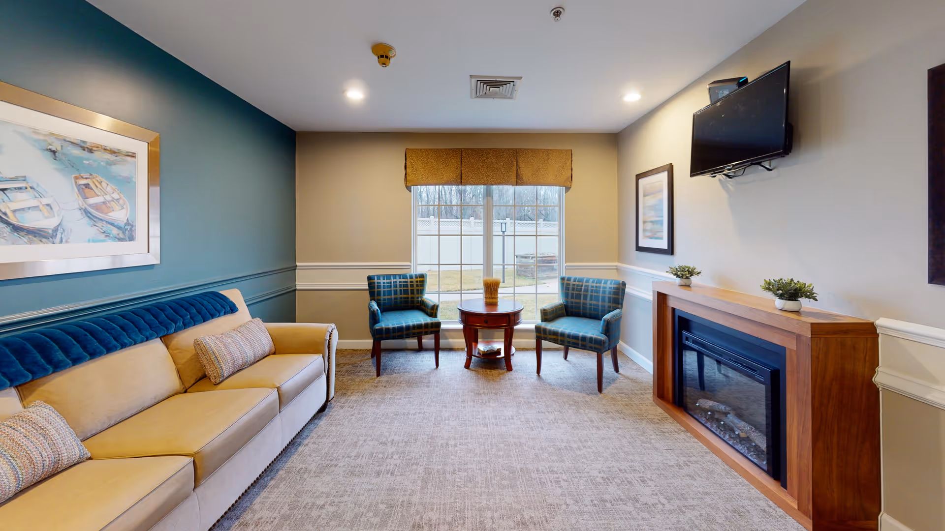 A cozy living room with a beige sofa and two patterned armchairs around a small wooden table in front of a large window. The room features a wall-mounted TV above a wooden electric fireplace, light beige walls with white trim, and a blue accent wall with a framed painting of boats. The floor is carpeted in a neutral tone.