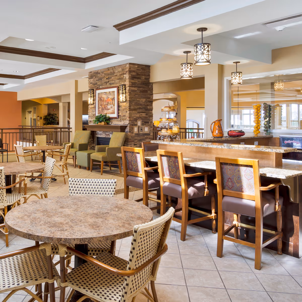 Bright communal dining and lounge area with round tables and wicker chairs, a stone-accented fireplace, and a bar with high stools.