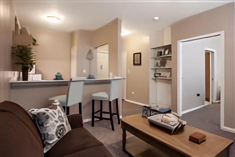 Interior view of a living room area with a brown couch and patterned pillows, a wooden coffee table with decorative items, two white bar stools at a counter, and an open doorway leading to a hallway with multiple doors.