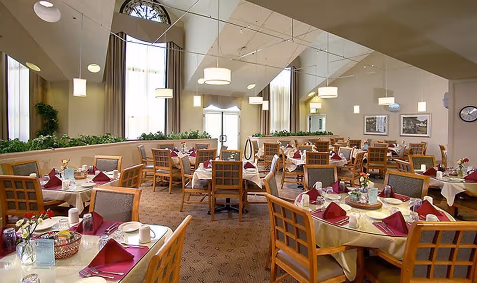 A spacious dining room with multiple round and rectangular tables set with white tablecloths, burgundy napkins, glassware, and small flower arrangements. The room has high ceilings with hanging pendant lights, large windows with curtains, and a mirrored wall reflecting the space.