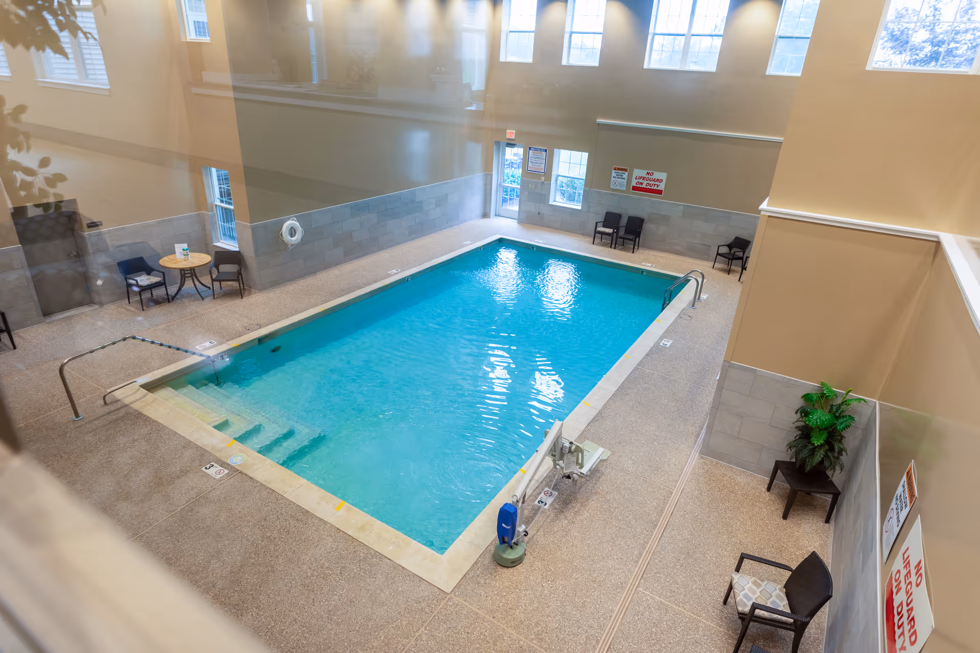 Indoor swimming pool in a bright enclosed room with seating, safety signs, and a pool lift.