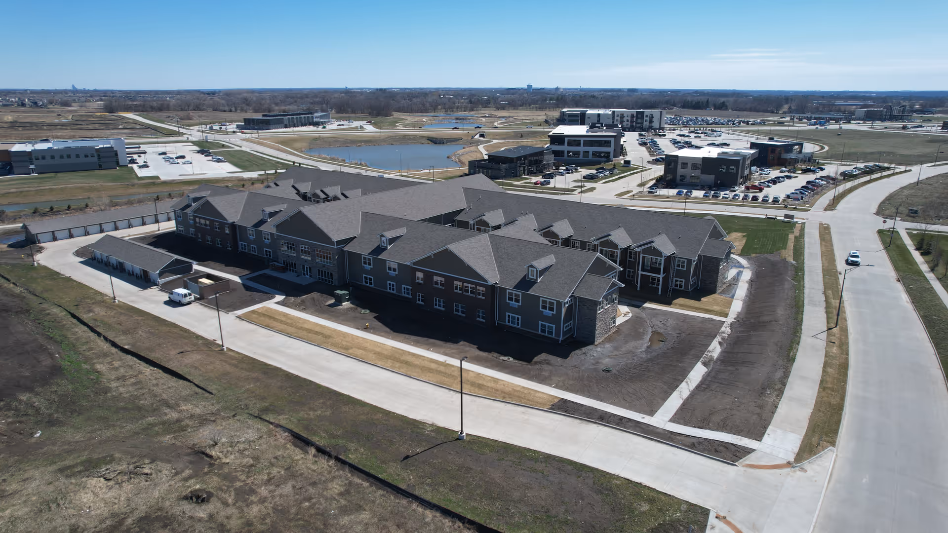 Aerial view of a large senior living facility building with multiple roof sections, surrounded by paved roads and parking areas. The landscape around the building is mostly bare soil and grass, with a pond and additional buildings visible in the background under a clear sky.