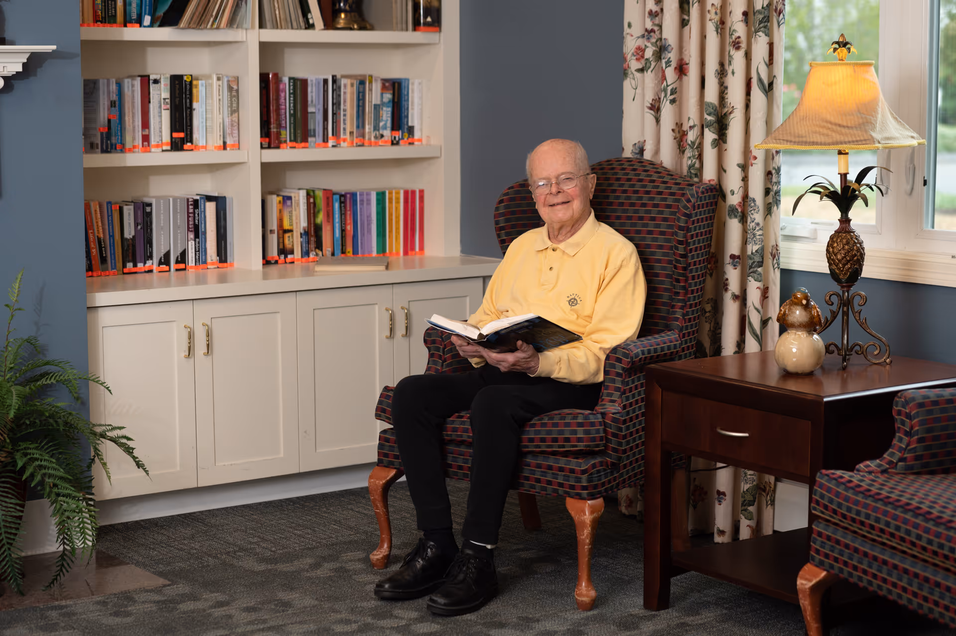 An elderly man sits in a patterned armchair reading a book in a cozy room with bookshelves, a side table, and a lamp.