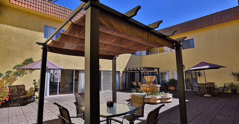 Sunny enclosed courtyard with a pergola, patio tables and chairs, umbrellas, and a central fountain surrounded by a two-story yellow building.