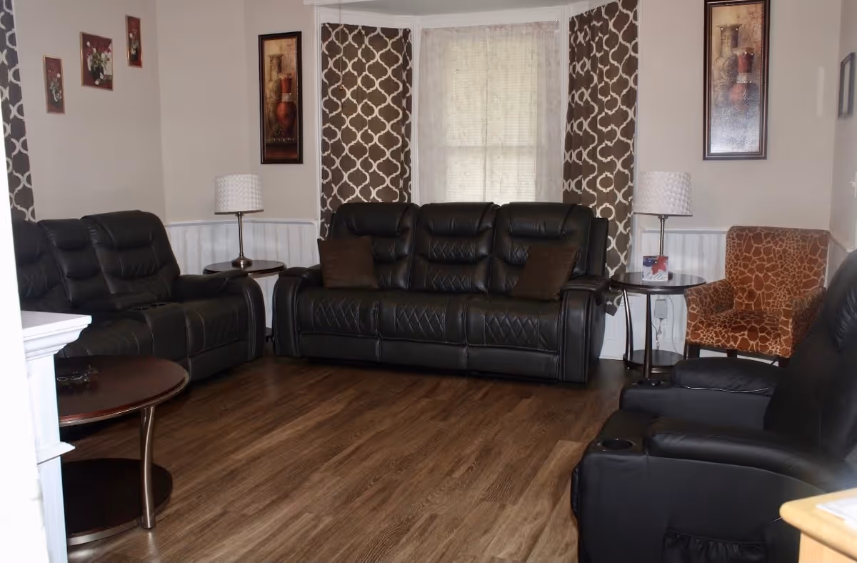 A living room with dark leather sofas and recliners arranged around a wooden floor. There are two side tables with lamps, a patterned armchair, and framed artwork on the walls. The window has patterned curtains and a sheer curtain in the center.