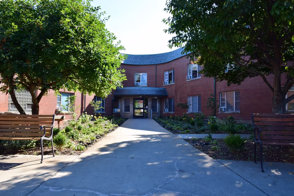 Outdoor view of the entrance to a red brick building with a curved roof section above the door. The pathway leading to the entrance is flanked by garden beds with plants and trees, and there are wooden benches on either side of the path.