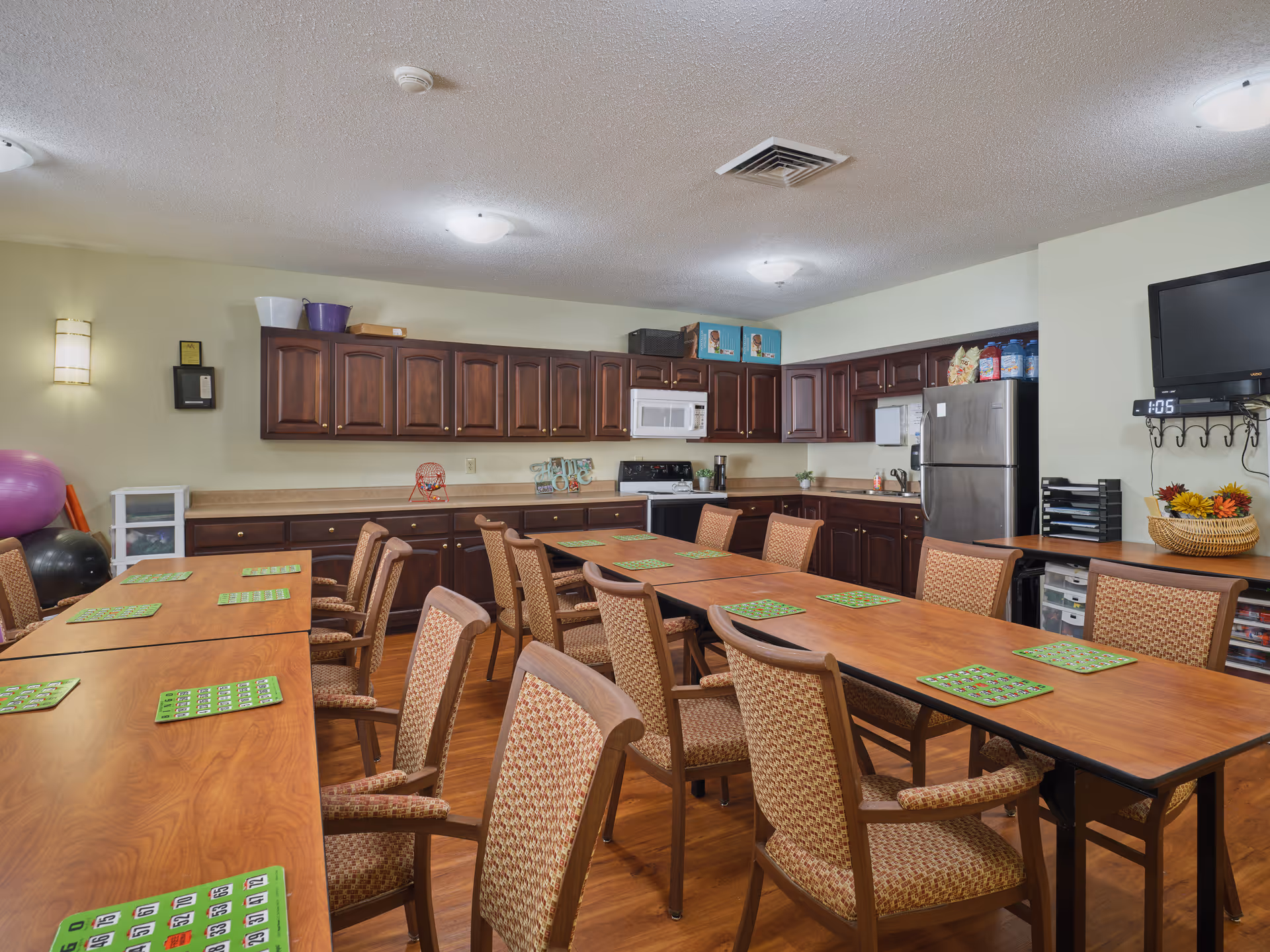A communal room with multiple wooden tables and cushioned chairs arranged for group activities. Each table has green bingo cards placed on it. The room features a kitchen area with dark wooden cabinets, a microwave, stove, and refrigerator. A wall-mounted TV is visible on the right side, along with some storage drawers and a basket with flowers. Exercise balls and other items are seen in the corner.