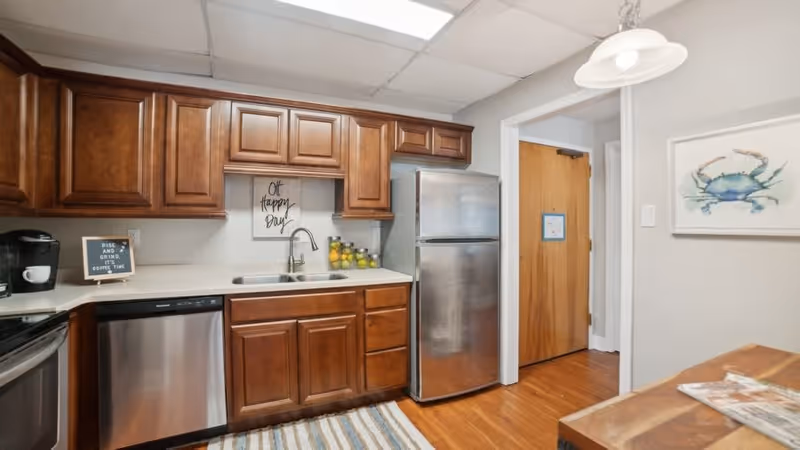 A kitchen area with wooden cabinets, a stainless steel refrigerator, dishwasher, and sink. There is a small sign on the counter that says 'Rise and Grind, It's Coffee Time' and a wall decoration above the sink that reads 'Oh Happy Day'. A wooden door and a framed picture of a crab are visible in the background.