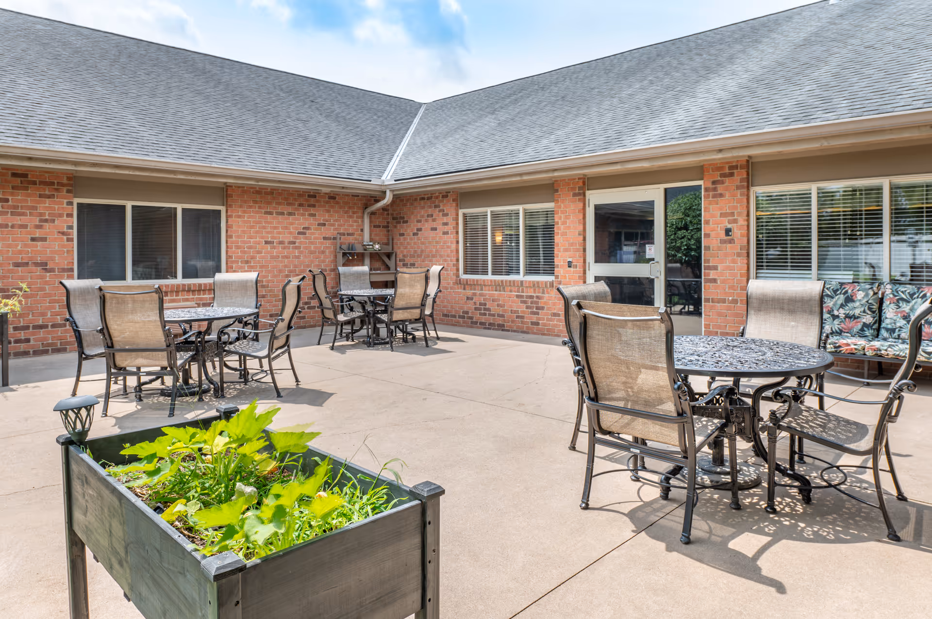 Outdoor courtyard patio with metal tables and chairs, a raised planter box, and a brick building entrance in the background.
