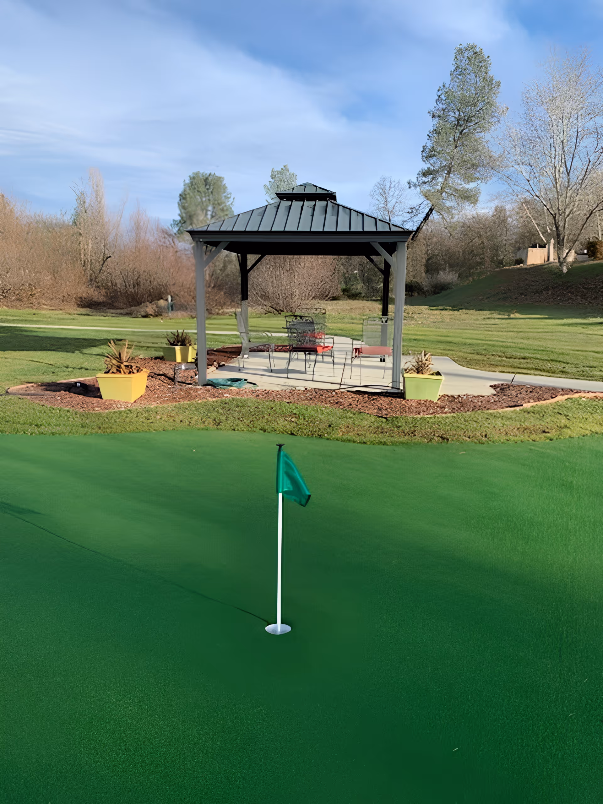 A small gazebo with chairs and tables beside a putting green and flag in a landscaped outdoor area.