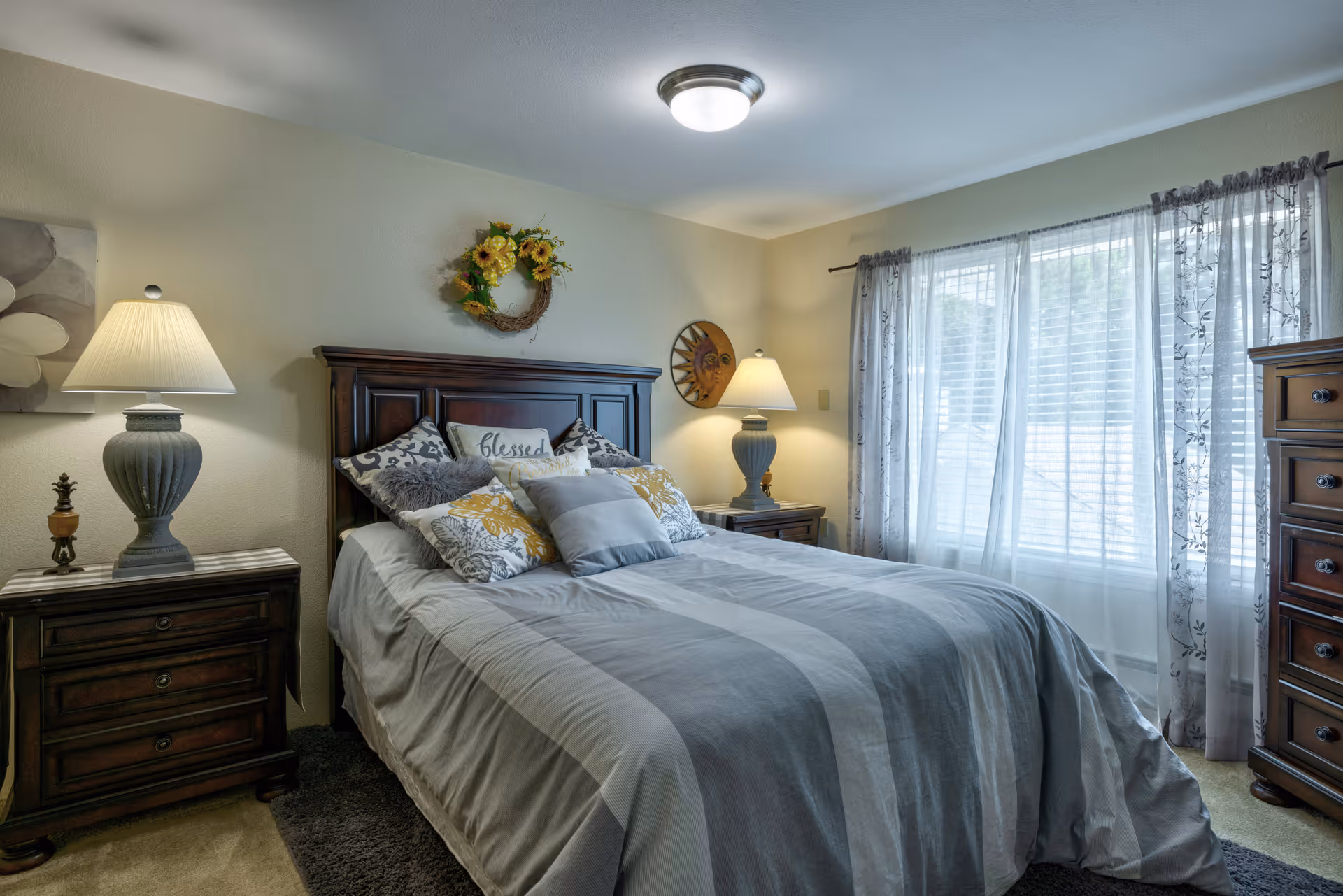 A cozy bedroom with a large bed covered in gray and white bedding and multiple decorative pillows. There are two wooden nightstands on either side of the bed, each with a lamp. A floral wreath hangs above the headboard, and sheer curtains cover a large window letting in natural light.