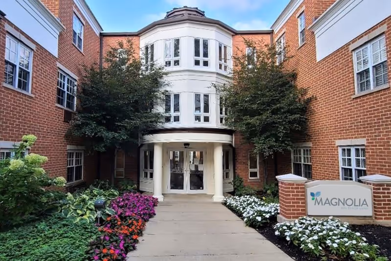 Entrance of a senior living facility with red brick walls, white framed windows, and a pathway lined with colorful flowers and green shrubs leading to double glass doors under a small portico supported by white columns. Two trees flank the entrance, and a sign on the right reads 'MAGNOLIA'.