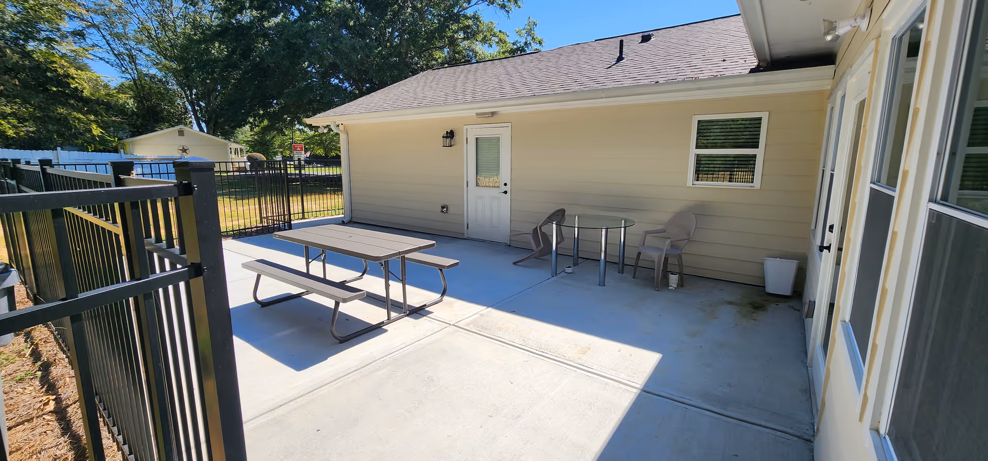Outdoor patio area with a picnic table and bench on the left side and a small round glass table with two chairs on the right side, adjacent to a beige building with a white door and window. The area is enclosed by a black metal fence, and there are trees and a small shed visible in the background under a clear blue sky.