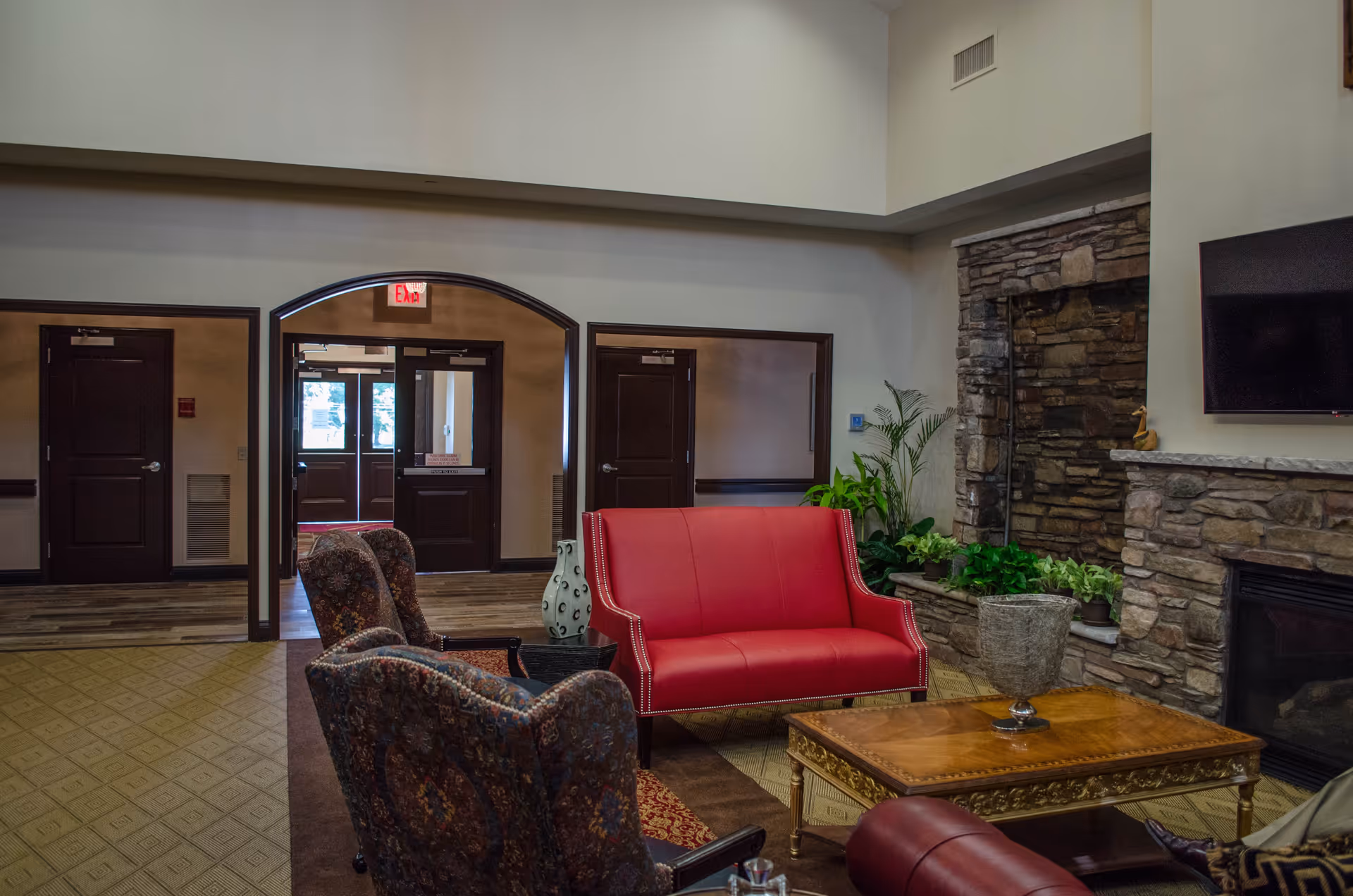 A cozy living room area in Meadow Falls Of Chagrin Valley featuring a red loveseat, two patterned armchairs, a wooden coffee table with a decorative vase, a stone fireplace, a mounted flat-screen TV, and several green plants. The room has high ceilings and an arched doorway leading to a hallway with dark wooden doors and an exit sign.