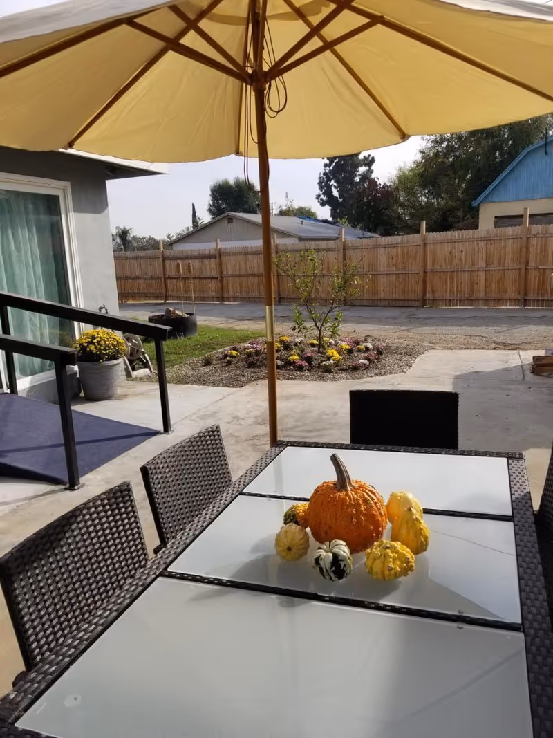 Outdoor patio with a glass-top table and wicker chairs under a large yellow umbrella, small pumpkins and gourds on the table, and a fenced yard with planters in the background.