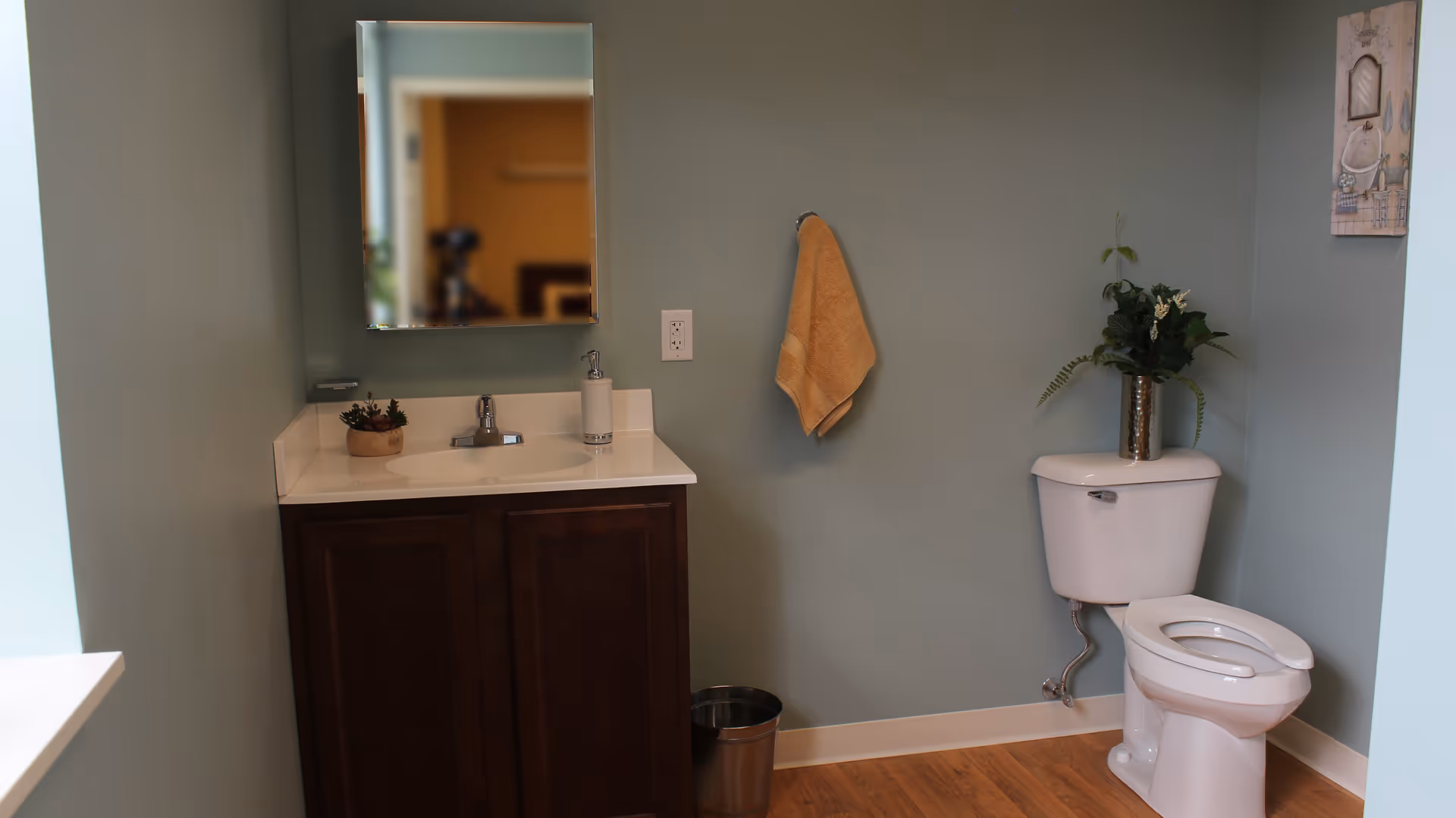 A bathroom with light blue walls, a white toilet with a silver vase holding green plants on top, a dark wood vanity with a white countertop and a sink, a soap dispenser, a small plant, a mirror above the sink, a beige towel hanging on a hook, and a small metal trash can on the wooden floor.