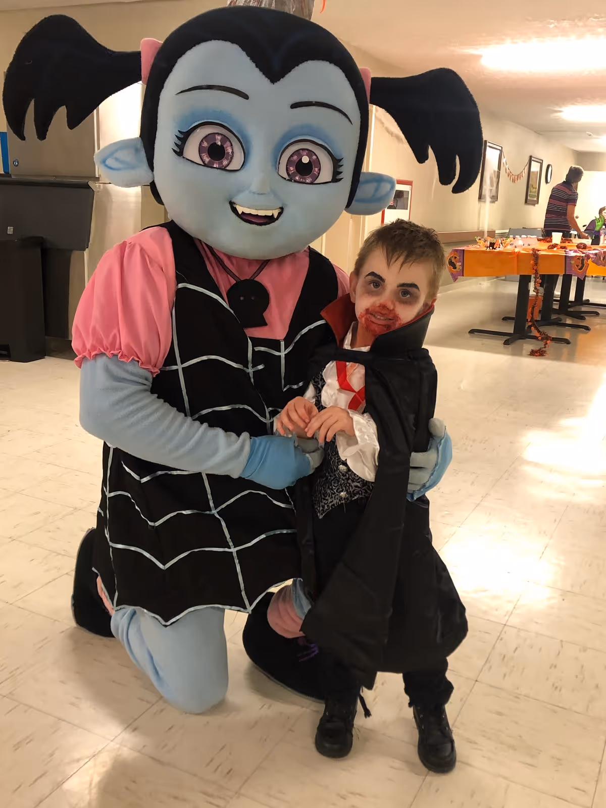 A person dressed in a large cartoon vampire costume with blue skin, black hair in pigtails, and a pink and black outfit kneels next to a young child dressed as a vampire with face paint and a black cape. They are indoors in a hallway decorated with Halloween-themed items on tables in the background.