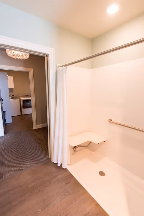 Accessible walk-in shower with a fold-down seat and grab bar, curtain open to a hallway showing a washer and dryer.