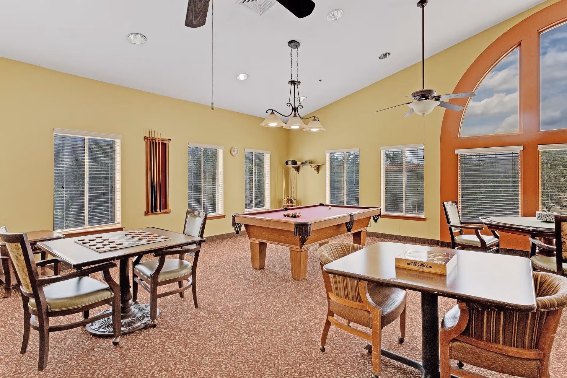 A bright game room with yellow walls and multiple windows. The room features a pool table in the center with billiard balls set up, a table with a checkers board and two chairs, and another table with two chairs and a book on it. Ceiling fans and hanging lights are visible, and there is a large arched window with blinds on the right side.