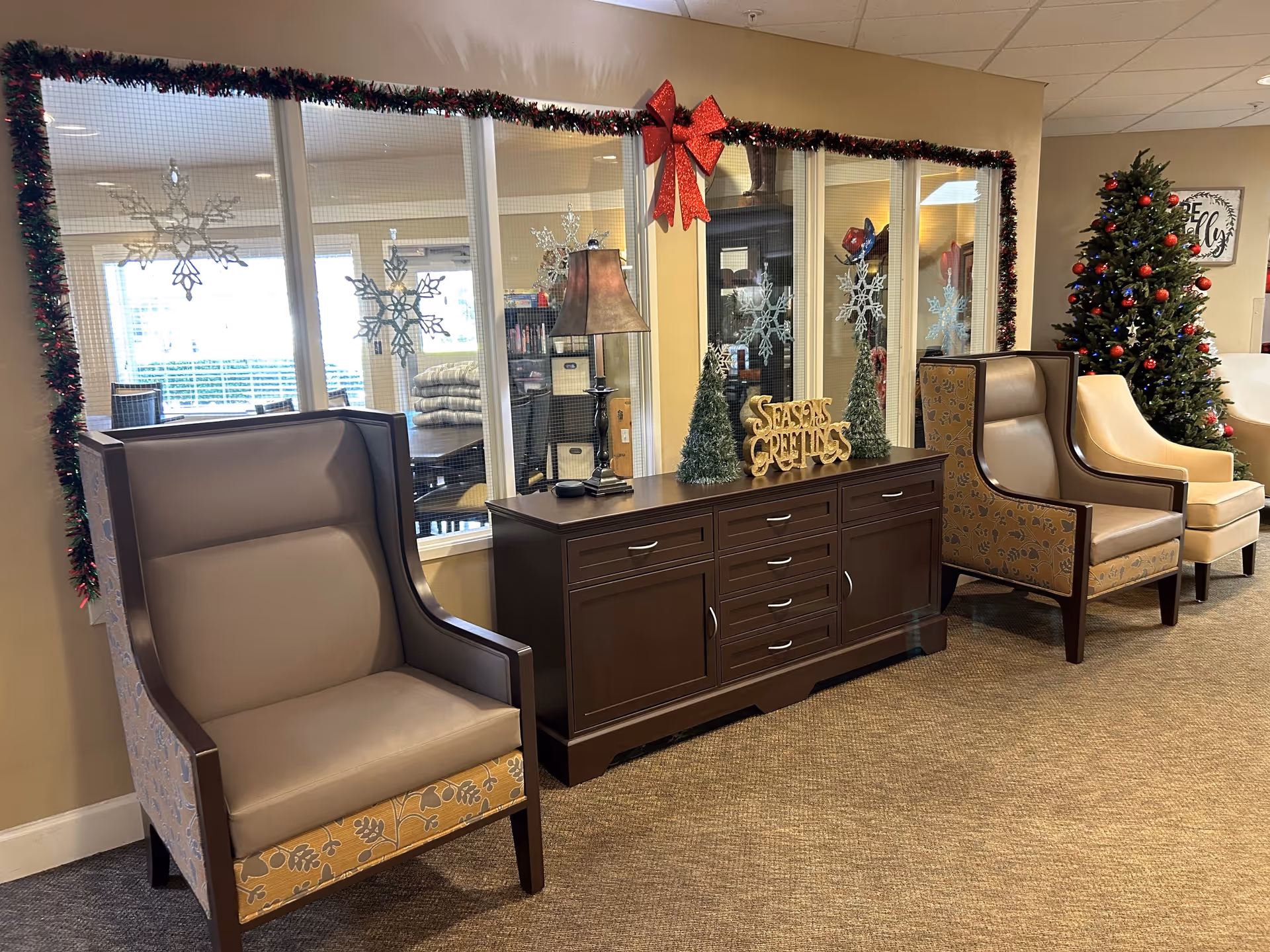 A cozy interior lounge area decorated for the holiday season with a Christmas tree adorned with red and blue ornaments, a dark wooden sideboard with a lamp, two small decorated Christmas trees, and a 'Seasons Greetings' sign. Two cushioned armchairs with patterned sides and a beige armchair are arranged along the wall. The wall behind features large windows decorated with garland, a red bow, and snowflake decals.