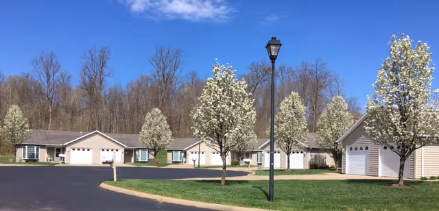 A row of single-story residential buildings with attached garages, surrounded by blooming trees and a paved driveway, under a clear blue sky.