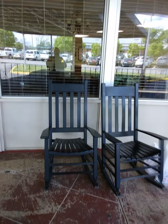 Two black wooden rocking chairs placed on a concrete porch in front of a window with blinds. The window reflects a parking lot with several cars and some greenery.