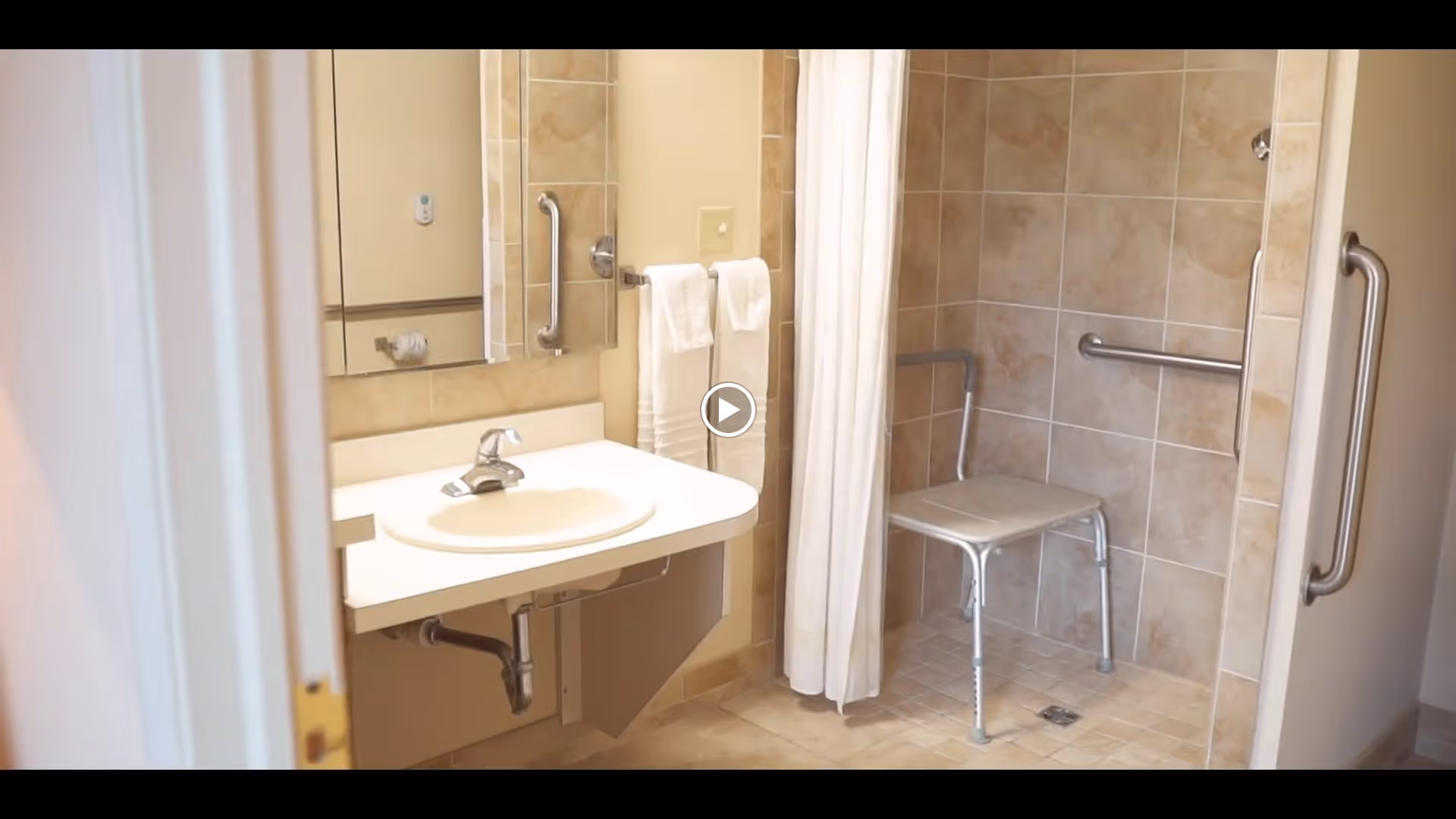 Accessible bathroom with a wall-mounted sink, a large mirror, grab bars, a towel rack with two white towels, and a walk-in tiled shower area featuring a shower chair and additional grab bars.